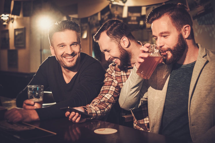 Cheerful old friends having fun with smartphone and drinking draft beer at bar counter in pub.
